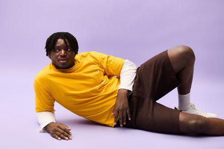 A young man in trendy clothes relaxes against a soft purple backdrop, radiating confidence.