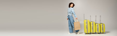 Beautiful young woman showcases trendy outfits while holding a shopping bag next to bright luggage.の写真素材