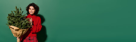 Stylish woman in a cozy sweater smiles with a holiday tree against a bright green background.の写真素材