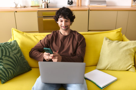 Handsome young man smiles while checking his smartphone and working on a laptop in a bright room.の写真素材