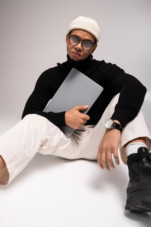 A handsome young man in stylish attire sits casually with a laptop, exuding confidence and flair.