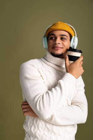 A stylish young man in a cozy sweater and hat smiles with a coffee cup, lost in music.の写真素材