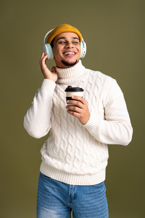 Stylish young man smiles while holding coffee cup and wearing cozy winter sweater and hat.