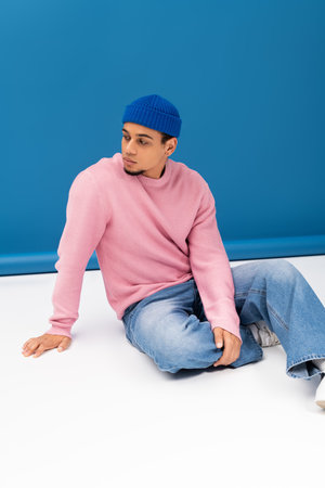 Handsome young man in trendy pink sweater and blue beanie sits on the floor, showcasing fashion.