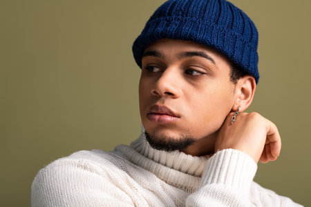 Young man in cozy sweater and beanie poses elegantly, expressing modern fashion sense and style.