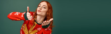 A cheerful young woman with red hair shows holiday spirit in a colorful Christmas sweater, bannerの写真素材