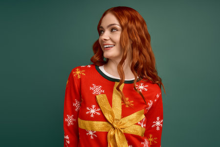 Young redhead woman smiles in a cozy Christmas sweater with a bow, celebrating winter holidays.