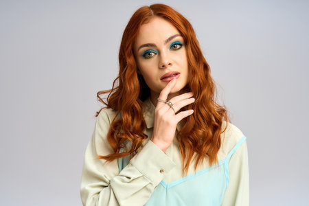 A redhead woman poses confidently against a grey background, showcasing bold eye makeup.