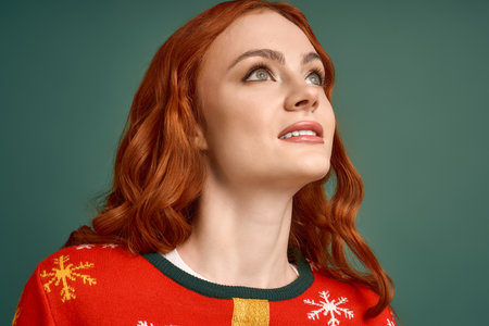 A joyful red-haired woman in a Christmas sweater poses against a green background.の写真素材