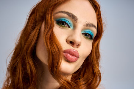 Young woman with red hair poses in studio, showcasing vibrant eye makeup and a fun expression.の写真素材