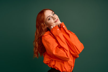 A confident redhead in an orange shirt stands against a green backdrop, showcasing autumn style.