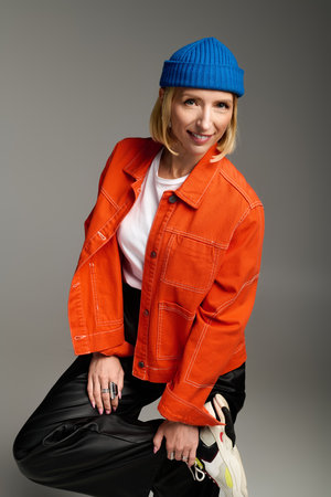 A fashionable blonde woman poses in a vibrant orange jacket and blue beanie, exuding joy.の写真素材