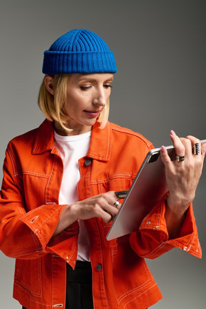 A fashionable blonde woman wears an orange jacket and blue beanie, focused on her tablet.の写真素材