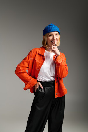 A trendy blonde woman poses playfully in a studio, wearing a blue beanie and orange jacket.の写真素材