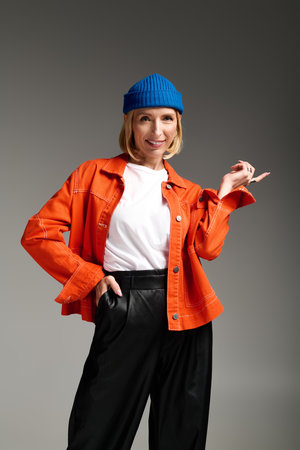 A fashionable blonde woman wearing a blue beanie and orange jacket poses confidently in a studio.の写真素材
