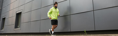 A young african american man jogs along a modern building's wall wearing a green hoodie and shorts,...