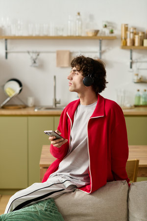 Handsome man in trendy attire listens to music on his phone in a cozy kitchen.
