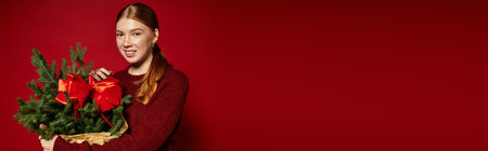 Young woman with red hair smiles while holding a pine tree against a vibrant red backdrop, bannerの写真素材