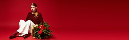 A young redhead poses with a small pine tree against a bold red background, embracing the holidays,...
