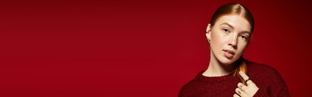 A confident young woman with radiant red hair poses in a knitted sweater against a red backdrop, bannerの写真素材