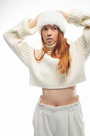 A young redhead poses gracefully in a cozy fur hat and soft sweater against white.の写真素材