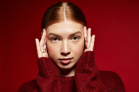 Red-haired young woman in a stylish winter sweater poses gracefully on a striking red background.の写真素材