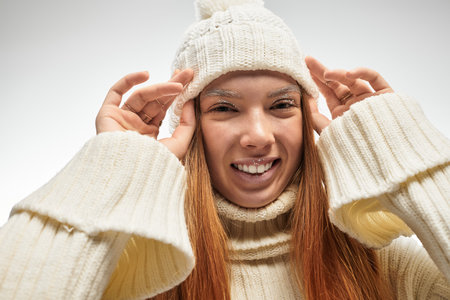 Redhead young woman poses playfully in a white sweater and hat, exuding winter charm.の写真素材