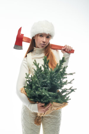 A young redhead in a fur hat and white sweater holds a small Christmas tree.の写真素材