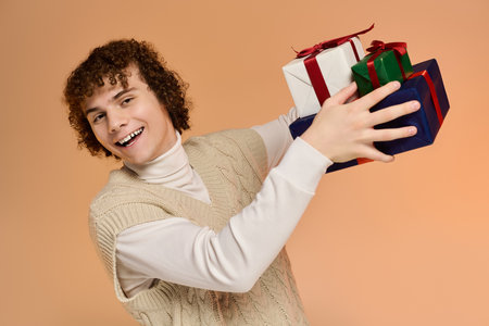 A cheerful teenage boy with curly hair holds colorful wrapped gifts in a cozy studio setting.