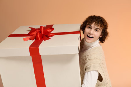 Curly-haired teenager in a cozy knitted vest joyfully holds a large holiday present.の写真素材
