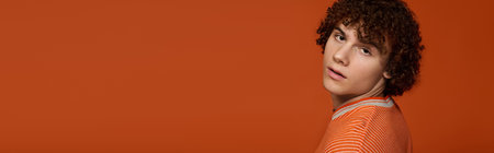 A confident young boy with curly hair stands in casual clothes against an orange background, bannerの写真素材