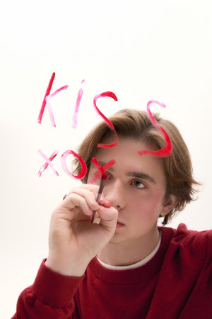 In a bright space, a young stylish man writes kiss with a marker while lost in thought.