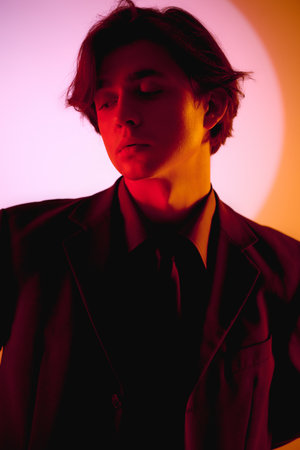 A fashionable young man stands confidently, illuminated by colorful lights in an artistic setting.