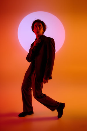A young man in a sleek outfit stands with one foot forward, exuding fashion dynamism in warm hues.