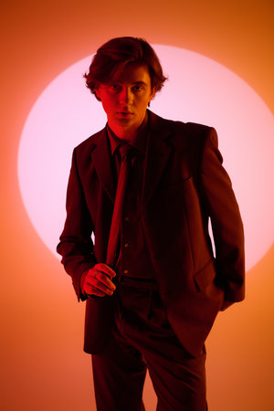 A young man in a stylish suit stands confidently before a bright, circular backdrop.