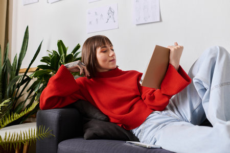 Young woman in a red sweater lounges at home, reading a notebook with plants nearby.の写真素材
