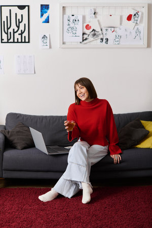Joyful young woman in a red sweater relaxes on her couch while working on her laptop at home.の写真素材