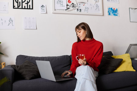 A young woman in a red sweater relaxes on her sofa, working on her laptop with tea.の写真素材