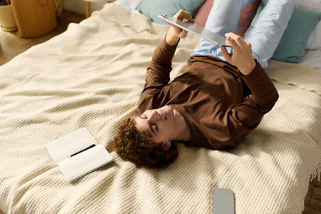 A teenage boy with curly hair lounges on his bed, using a tablet while at home.の写真素材