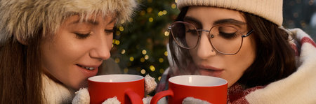 Two women share warm drinks while wrapped in cozy attire, surrounded by holiday cheer.の写真素材