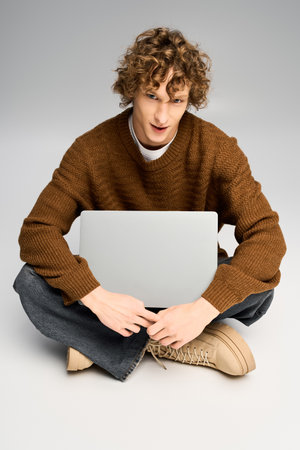 Handsome young man in cozy knit sweater and stylish pants sits cross legged with laptop, smiling.の写真素材