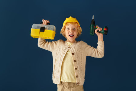 A cheerful blonde boy with wavy hair holds a tool kit and a drill in a studio setting.の写真素材