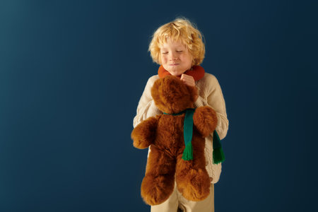 A happy blonde boy with wavy hair enjoys holding his teddy bear in a cozy studio setting.の写真素材