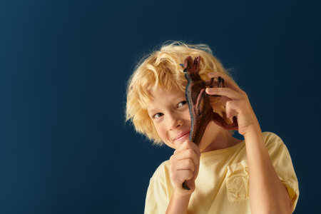 A carefree blonde boy with wavy hair smiles while holding a dinosaur toy in a studio setting.の写真素材