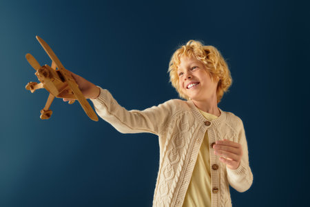 A cheerful blonde boy holds a wooden airplane while smiling in a blue studio.の写真素材