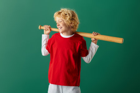 A carefree blonde boy in red clothes playfully poses with a bat, smiling against the green backdrop.