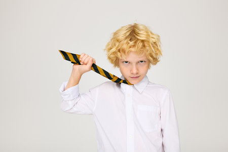 A confident blonde boy in uniform holds his striped tie proudly, exuding joy.の写真素材