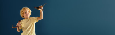 A carefree blonde boy with wavy hair enjoys playing with his dinosaur toys in a studio, bannerの写真素材