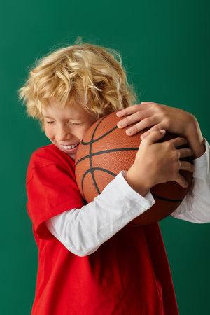 A happy blonde boy in red clothes smiles with a basketball against a green backdrop.の写真素材