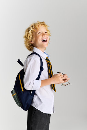 Blonde boy in a school uniform beams with happiness, holding glasses and backpack in studio light.の写真素材